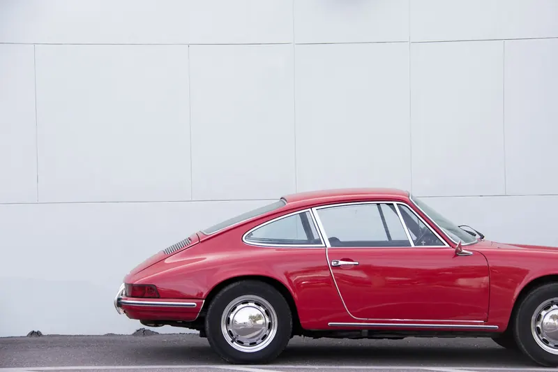 Red early Porsche 911 coupé, seen from the side against a plain white wall.
