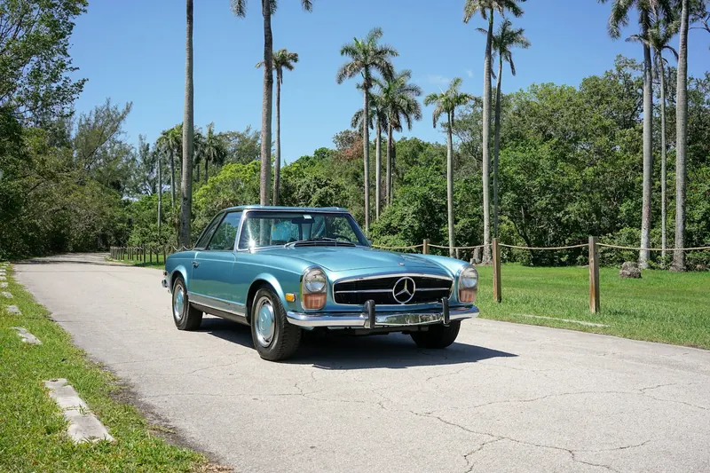 Pale blue Mercedes-Benz 280 SL with its hardtop on, on a lane lined with palm trees.