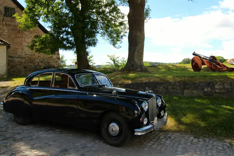 Black Jaguar Mark VIII saloon on cobbles, beside an old brick building and a tree.