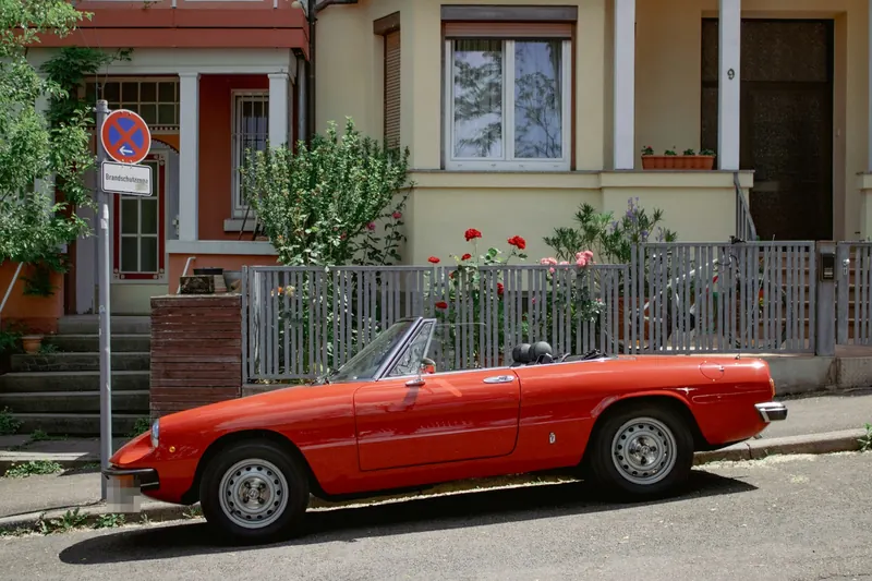 Red Alfa Romeo Spider, roof folded, outside a row of painted houses.