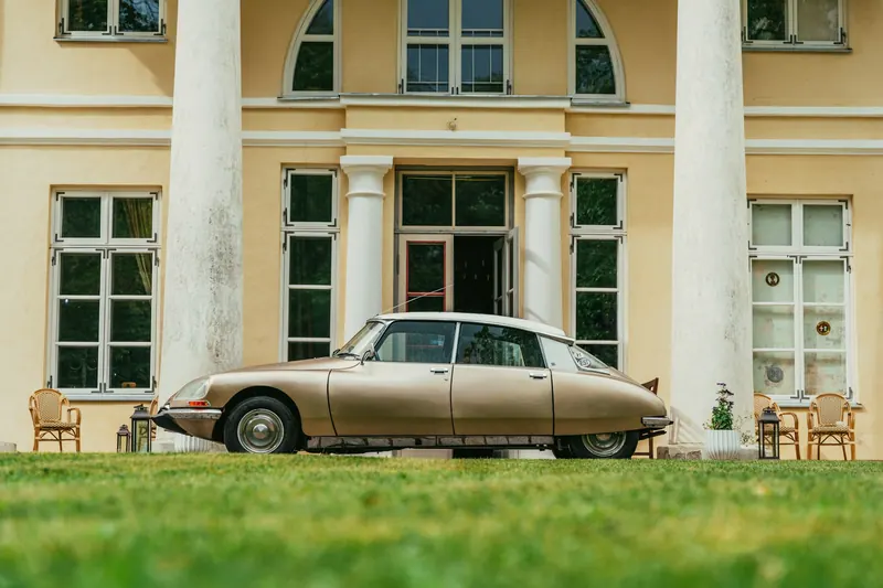 Gold Citroën DS with a white roof, side view, on a lawn in front of a pale yellow house with columns.