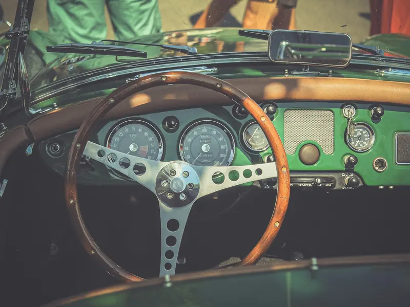 Inside a green open car: a wood-rimmed steering wheel, round dials and a brown leather roll along the dash.